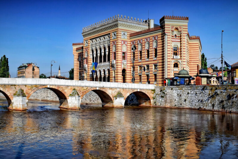 Sarajevo City Hall with a bridge in the foreground, reflecting in the river under a clear blue sky.