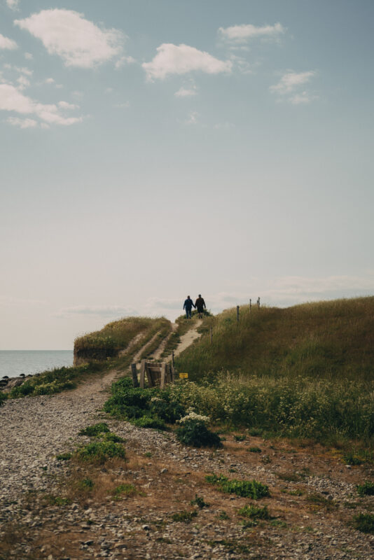 Two people walking along a path above the waves, surrounded by grassy hills and a serene sky.