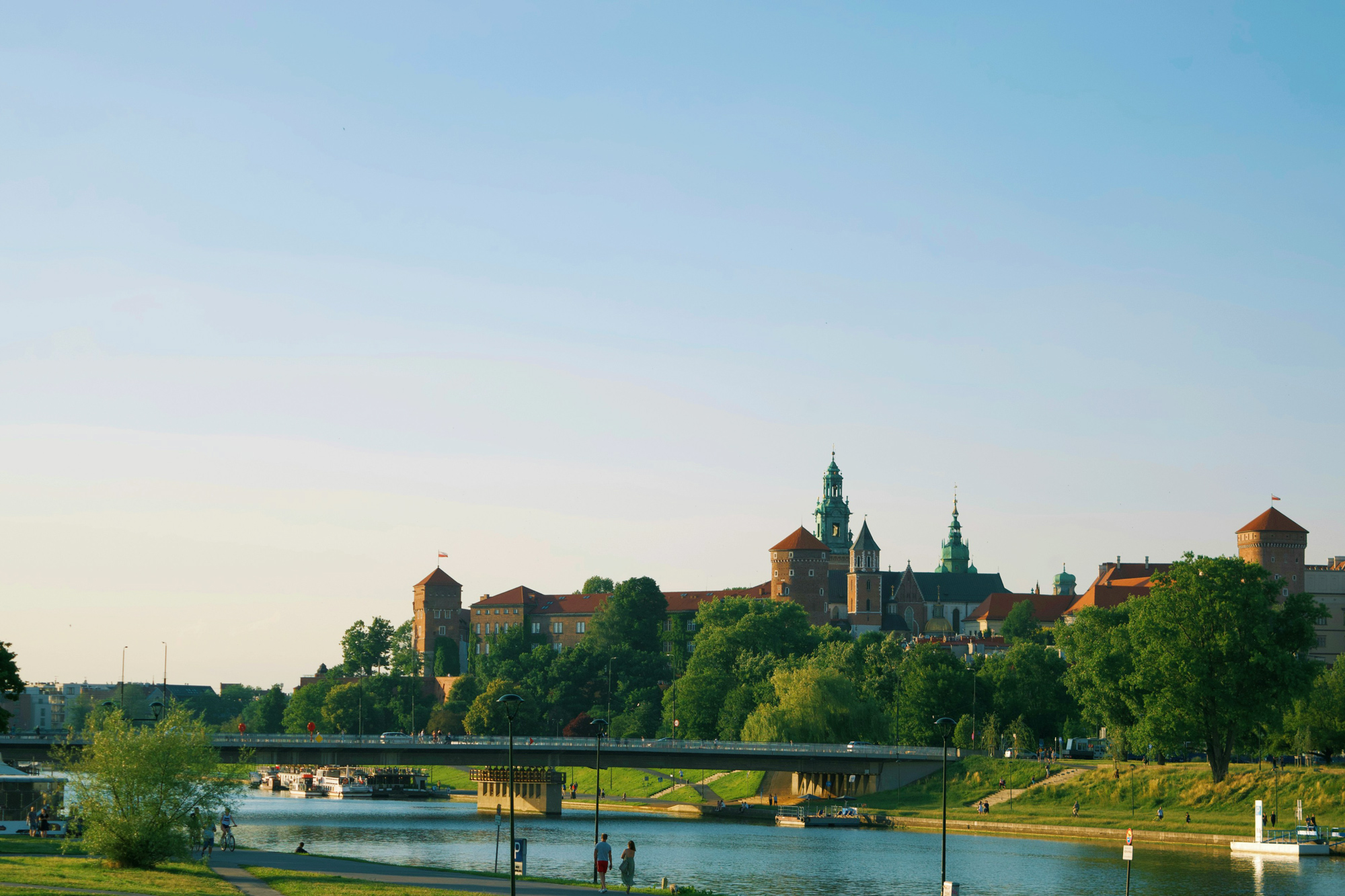 View of a river with a bridge, leading to a castle and towers on a hill under a clear blue sky.