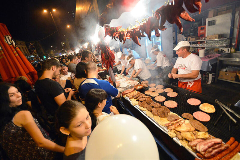 Crowd enjoying a street food festival with vendors grilling various meats at night, smoke rising from the grill.