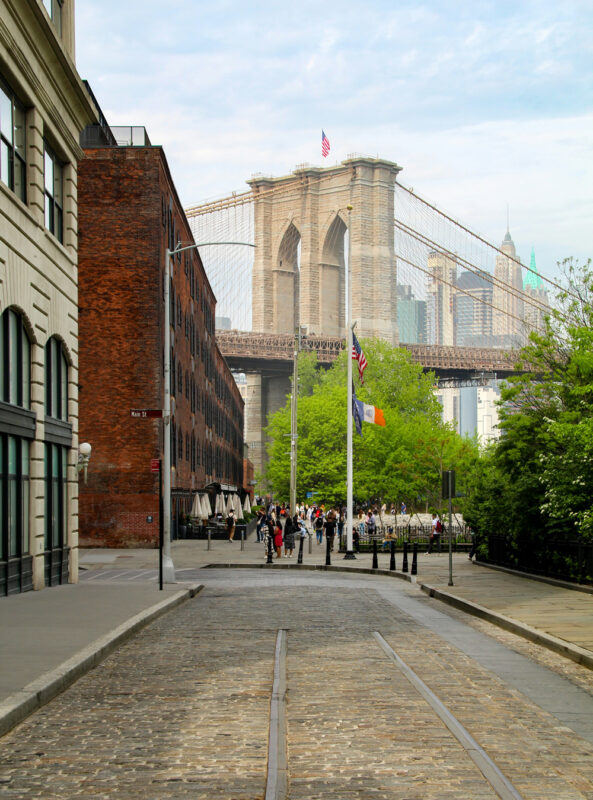Cobblestone street leading to Brooklyn Bridge, with people walking and greenery in the foreground.