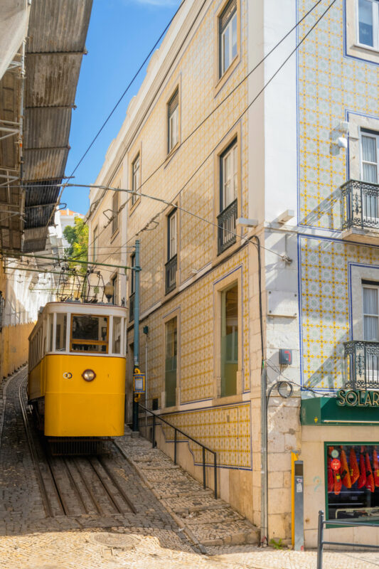 Yellow tram ascending a steep cobblestone street, flanked by patterned buildings in Lisbon.