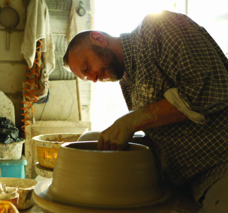 A man with a beard shapes clay on a pottery wheel in a sunlit workshop, surrounded by tools and pottery.
