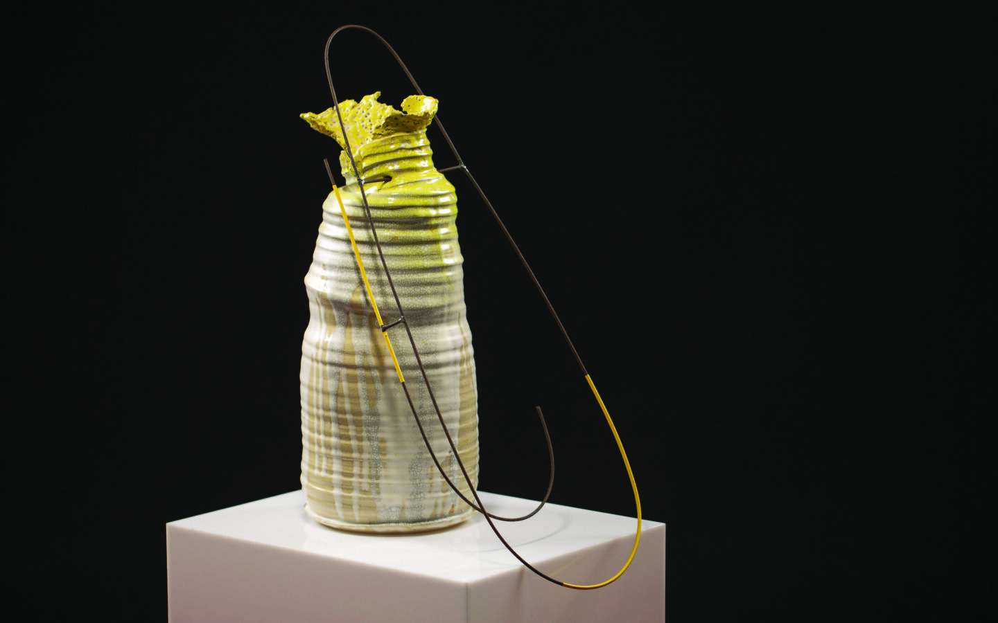 Ceramic vase with a textured surface, topped with a green leaf-like sculpture, displayed on a white pedestal against a bla...