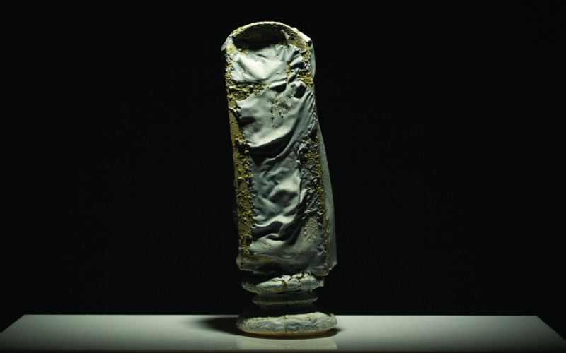 Abstract sculpture with a textured, dark surface, displayed on a white pedestal against a black background.