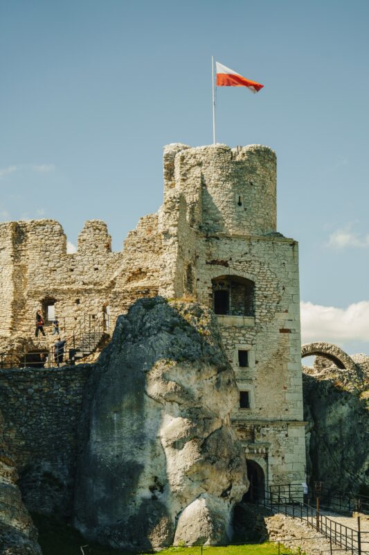 Ruins of a stone castle with a Polish flag atop, surrounded by rocky terrain and a clear blue sky.