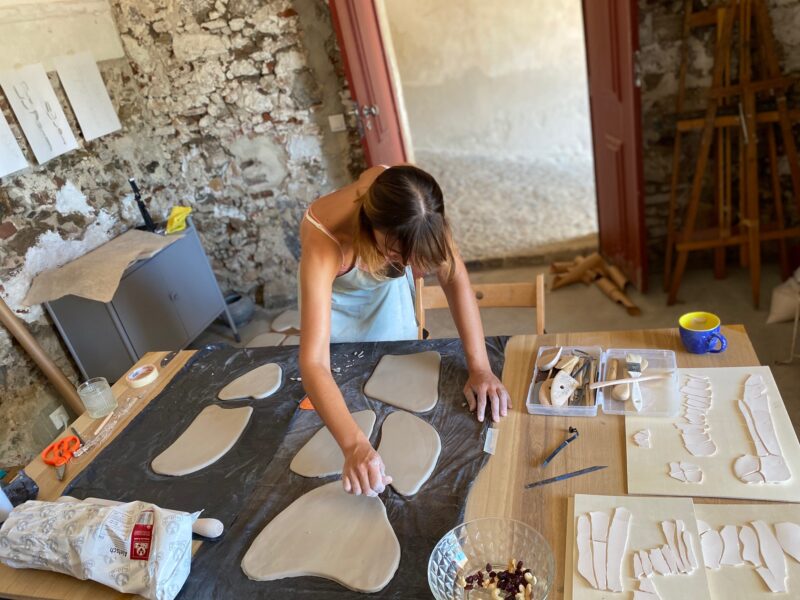 Artist shaping clay pieces on a table in a rustic studio, with tools and materials scattered around.
