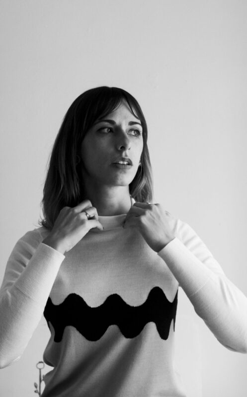 Woman adjusting her sweater with a wave pattern, standing in a softly lit room with plates in the background.