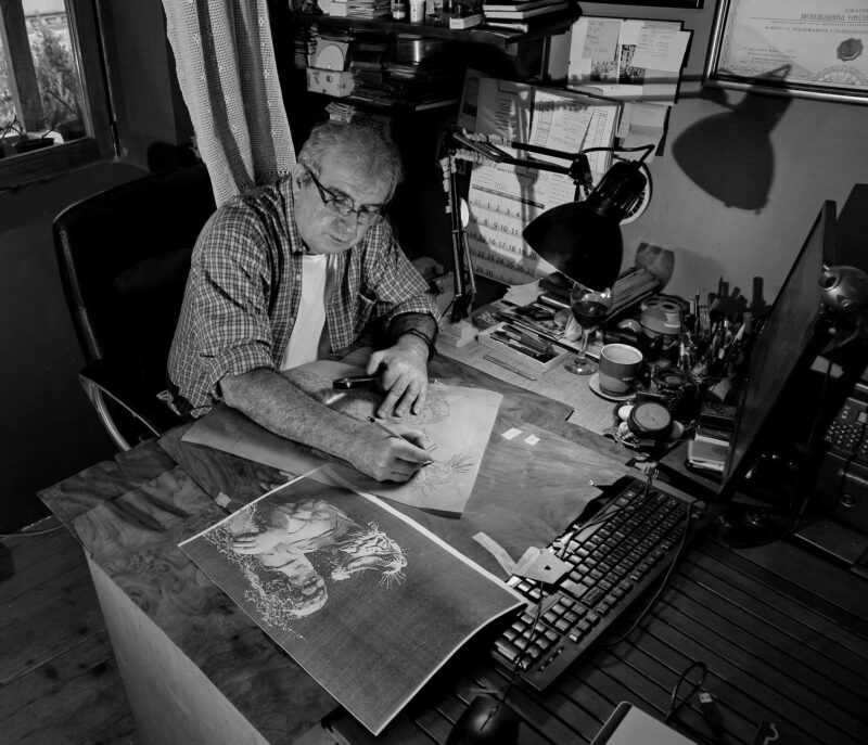 An older man with glasses sketches at a cluttered desk, surrounded by art supplies and papers in a black-and-white setting.