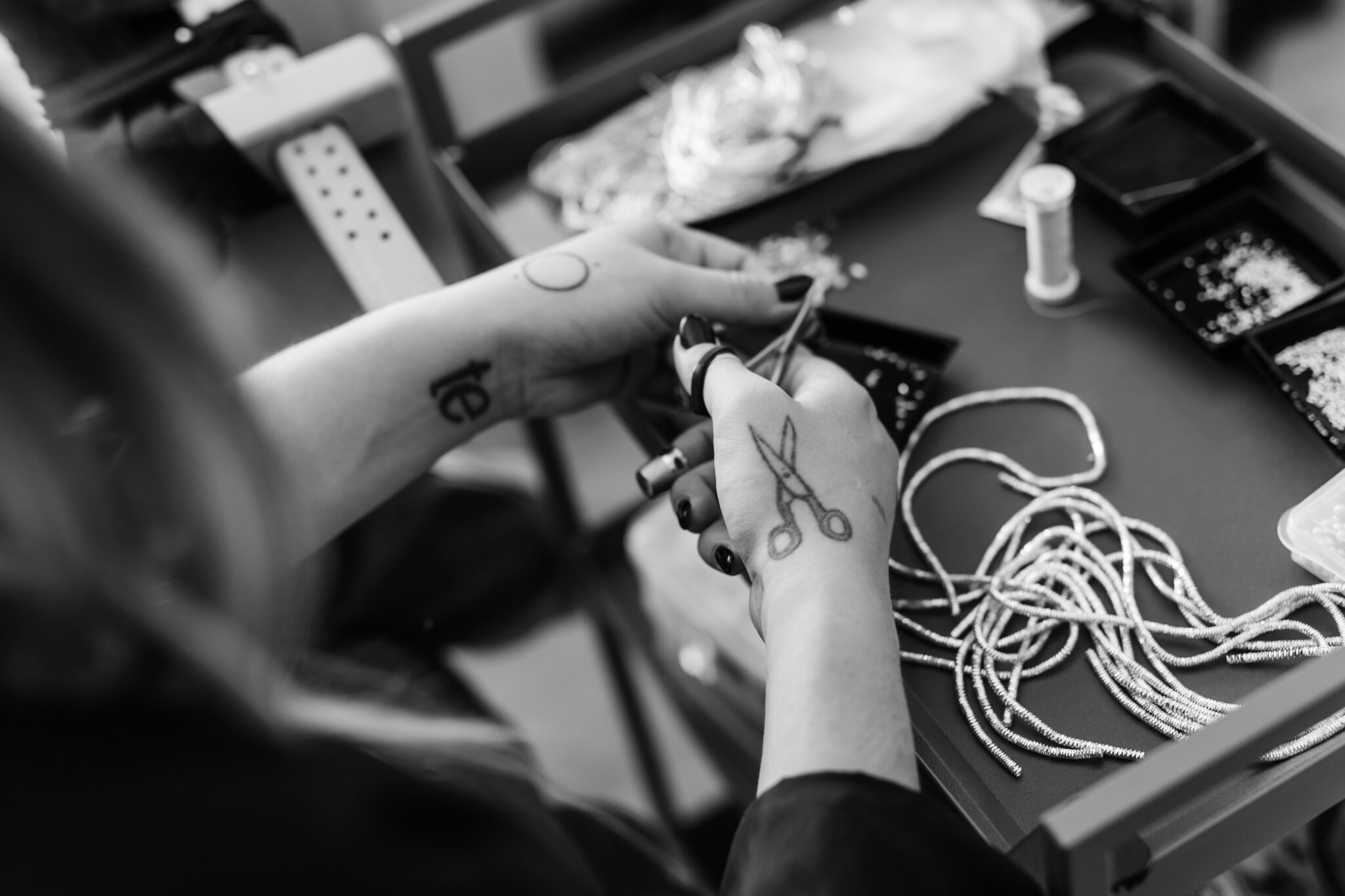 Hands with tattoos cutting threads, surrounded by beads and tools on a work surface.