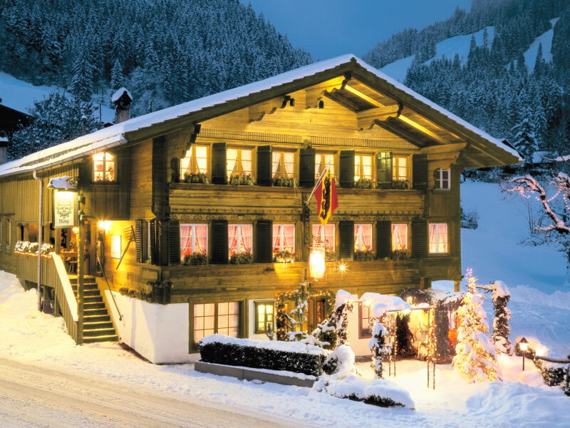 Charming wooden chalet in a snowy landscape, warmly lit with festive decorations and a Swiss flag.