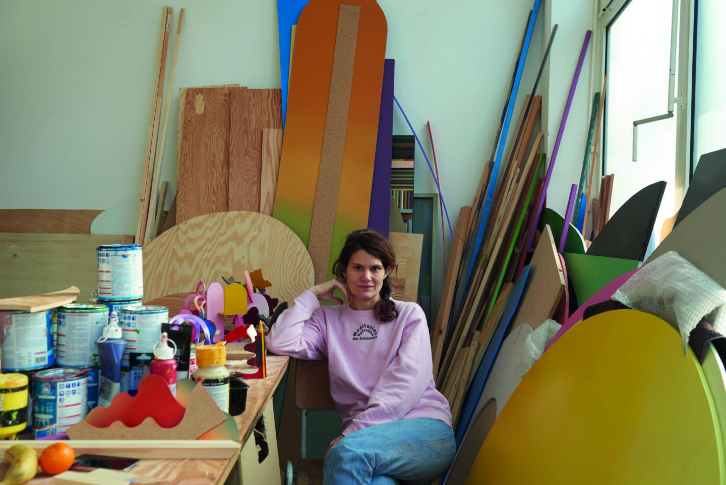 A woman in a pink sweater sits in an art studio surrounded by colorful materials and paint cans.
