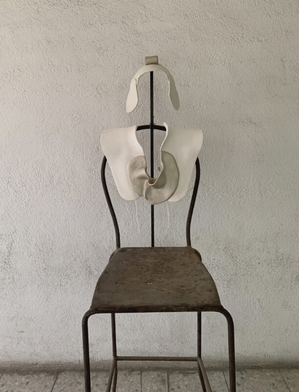 Sculptural piece resembling a torso mounted on a metal chair against a textured white wall.
