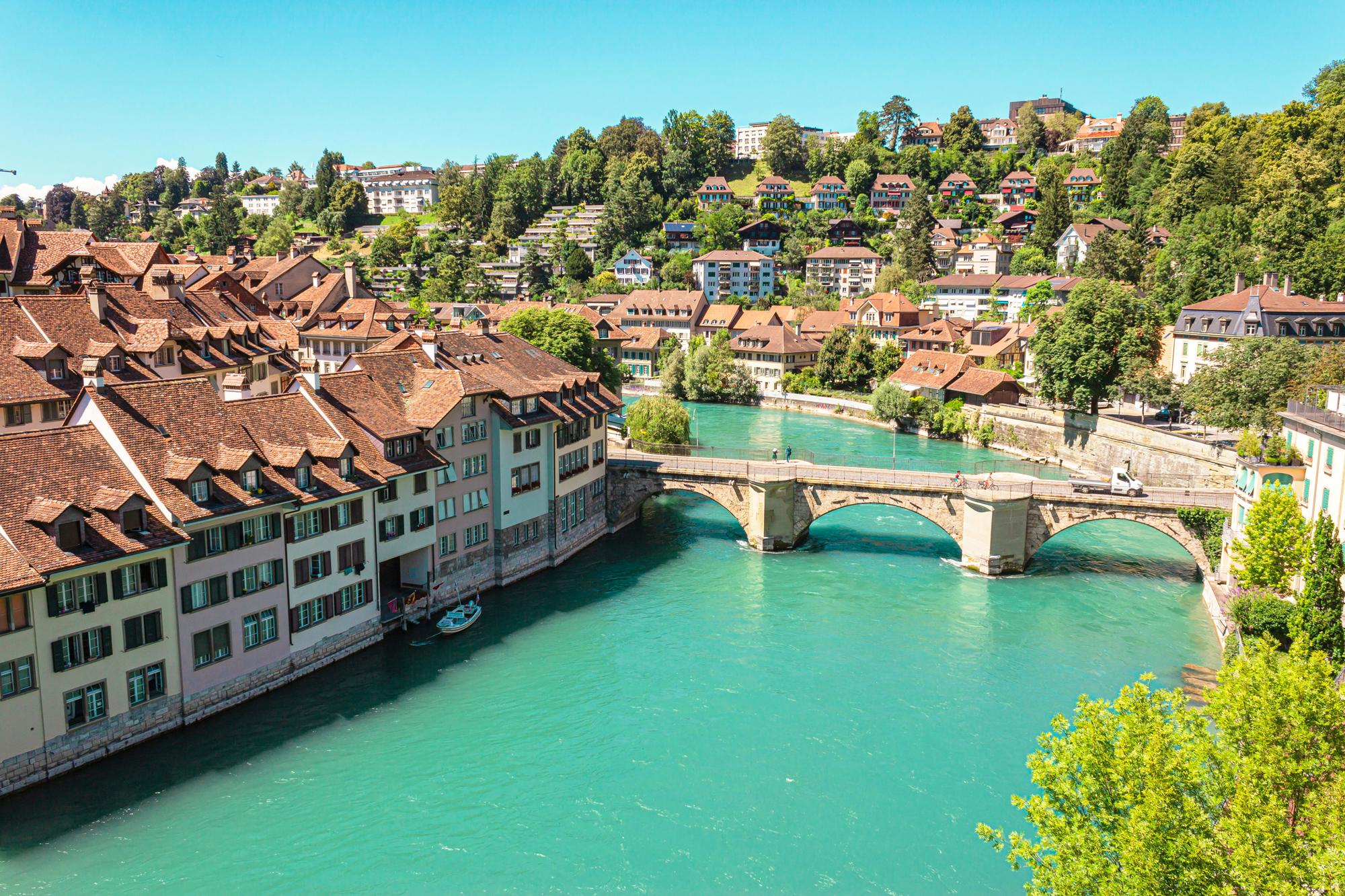 View of a turquoise river flowing through a city with traditional buildings and a stone bridge, surrounded by green hills.