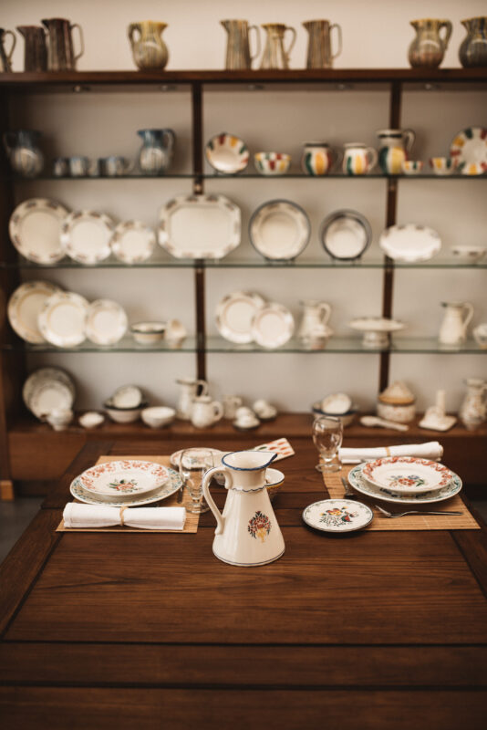 Ceramic table setting with a decorative pitcher, plates, and glasses, displayed against a shelf of various ceramics.