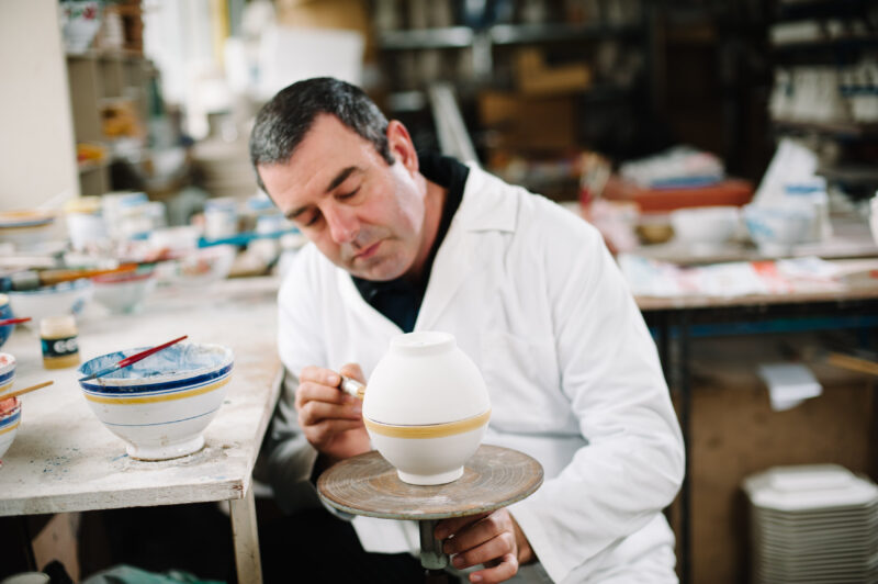 A man in a white coat carefully paints a decorative line on a ceramic vase in a pottery studio.