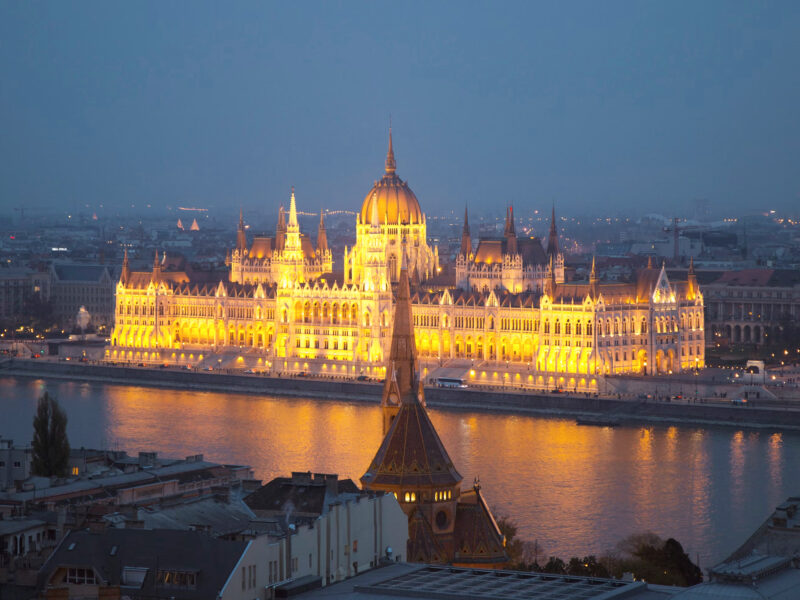 Budapest's Parliament building illuminated at dusk, reflecting in the Danube River, with a tower in the foreground.