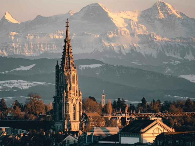 Historic tower with spire in foreground, snow-capped mountains in background, bathed in warm sunset light.