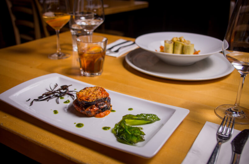 Elegant dish featuring layered ingredients with a crispy topping, garnished with greens and sauce, set on a wooden table.