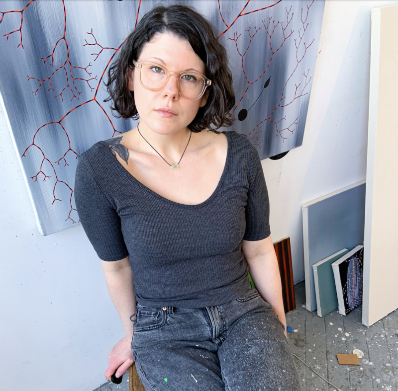 Woman with short dark hair and glasses, wearing a gray top, seated in an art studio with abstract paintings in the backgro...