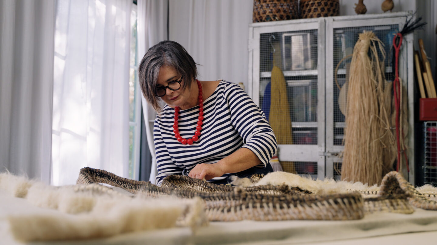 Maria Pratas, wearing a striped shirt and red necklace, works on textile art in her studio, surrounded by materials.