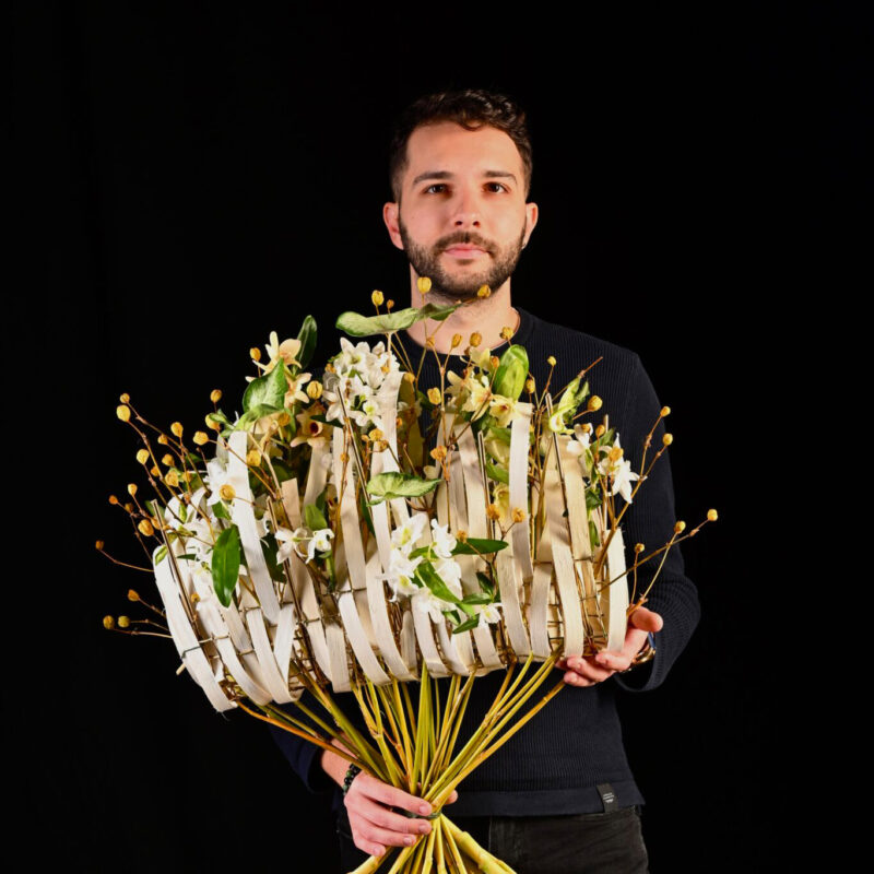 Man holding a large bouquet of flowers and branches against a black background.