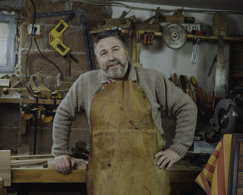 Man in a workshop wearing an apron, smiling while leaning on a wooden workbench surrounded by tools.