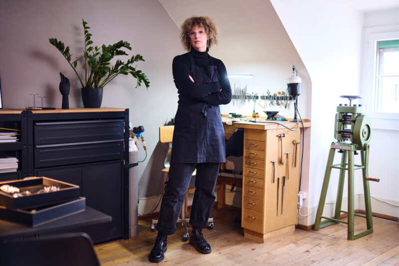 Portrait of Sarah Küffer in her workshop, wearing a black apron, standing confidently with arms crossed beside work tools.