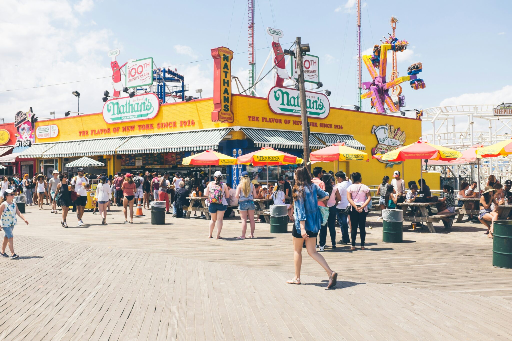 Crowded boardwalk scene at Nathan's Famous, featuring bright yellow building and colorful umbrellas on a sunny day.
