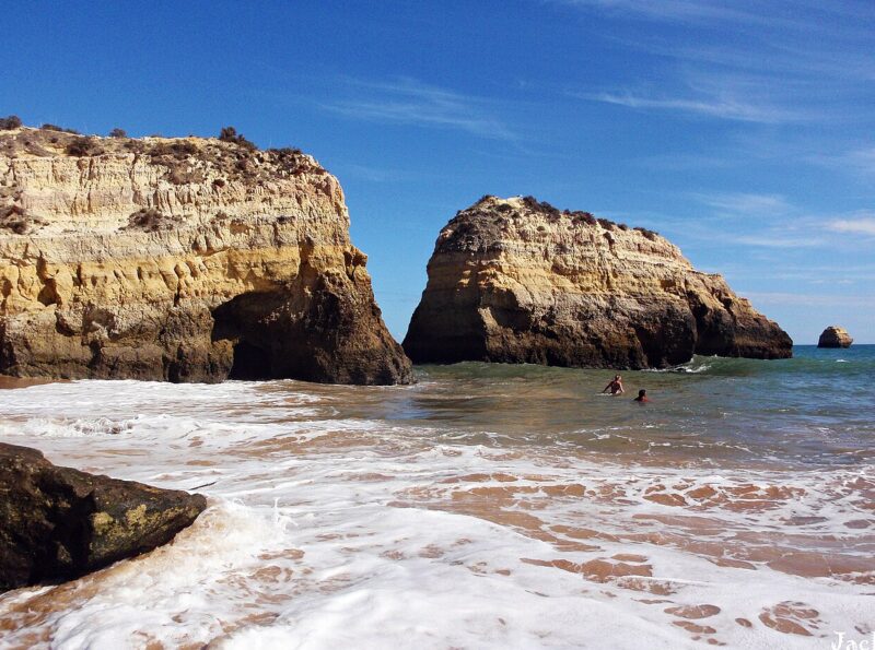Rocky cliffs rise above a sandy beach with waves crashing, while two people swim in the clear water under a blue sky.