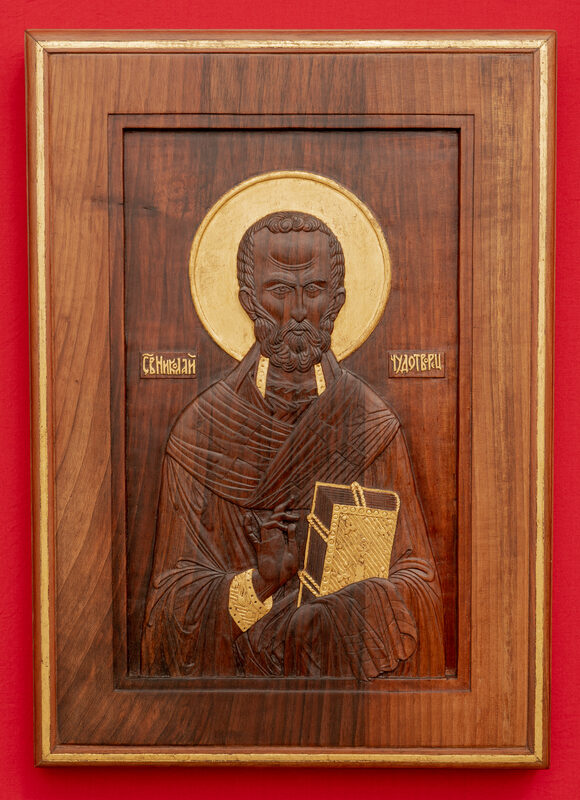 Carved wooden icon of Saint Nicholas, featuring a golden halo and holding a book, set against a red background.