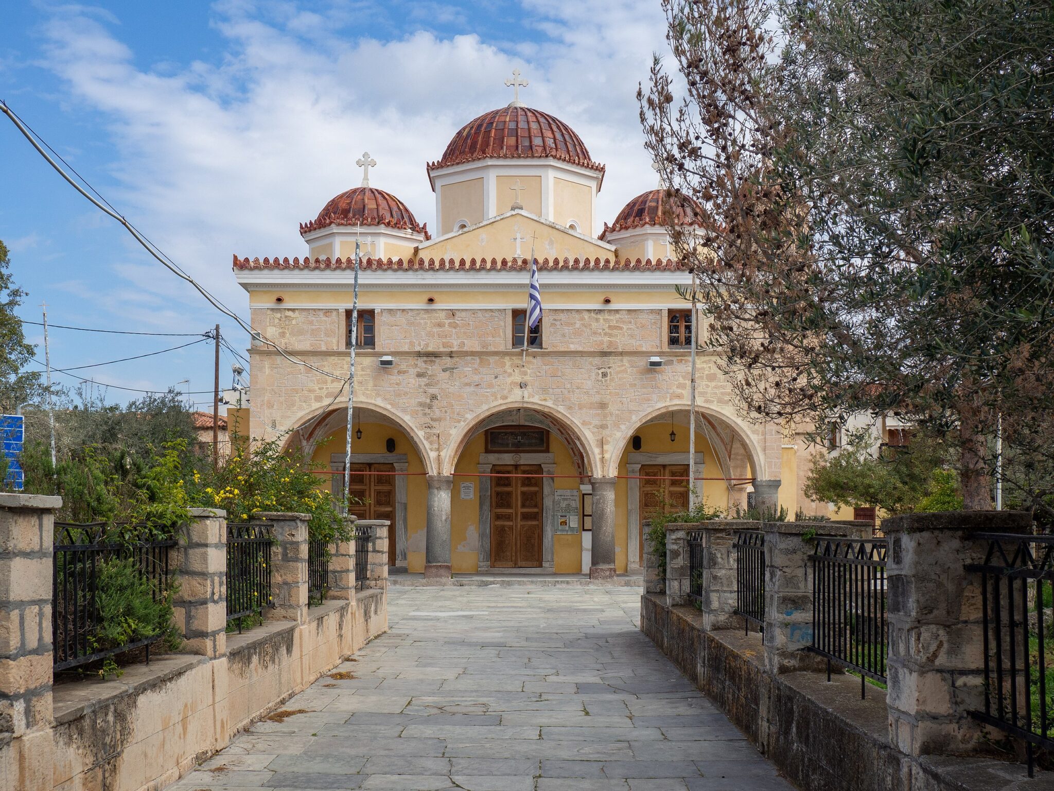 Metropolitan Church of the Dormition of the Theotokos in Aegina, featuring domes and a stone facade, under a cloudy sky.