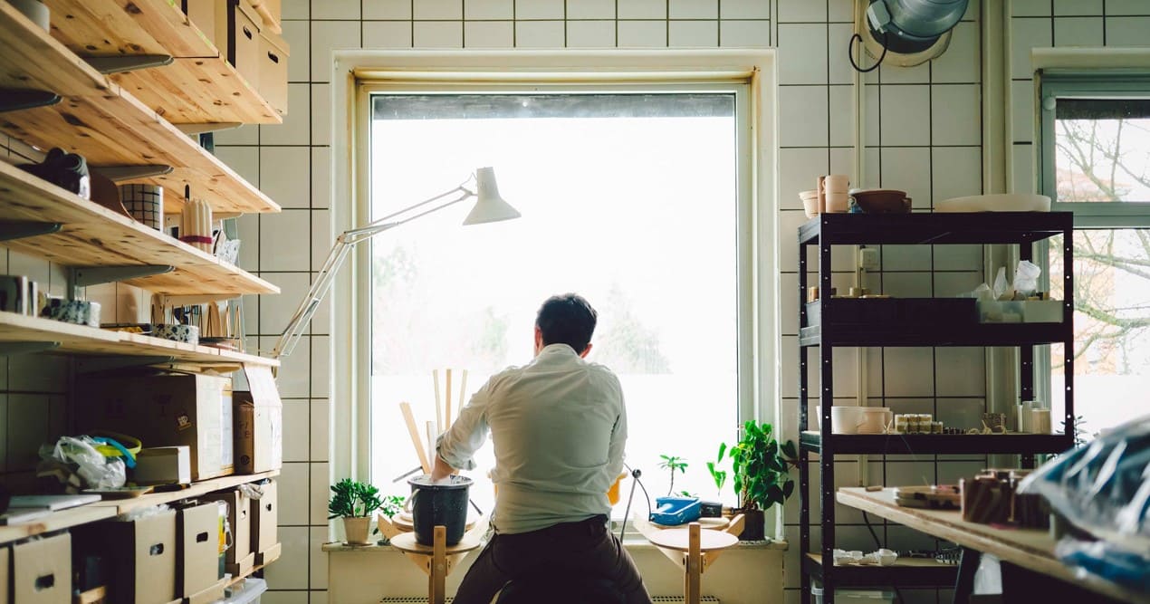 Oya in his studio, seated with his back to the camera, surrounded by plants and art supplies, looking out a large window.