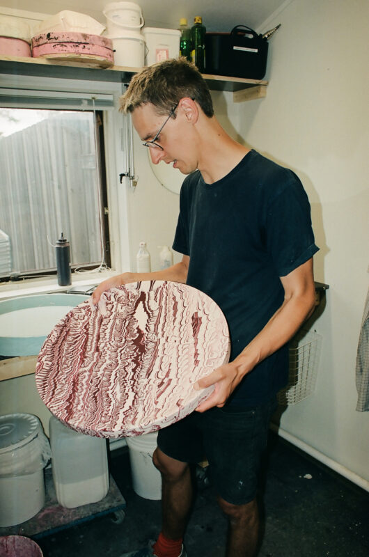 A person holds a large, marbled platter with pink and white patterns, standing in a workshop with various tools and materi...