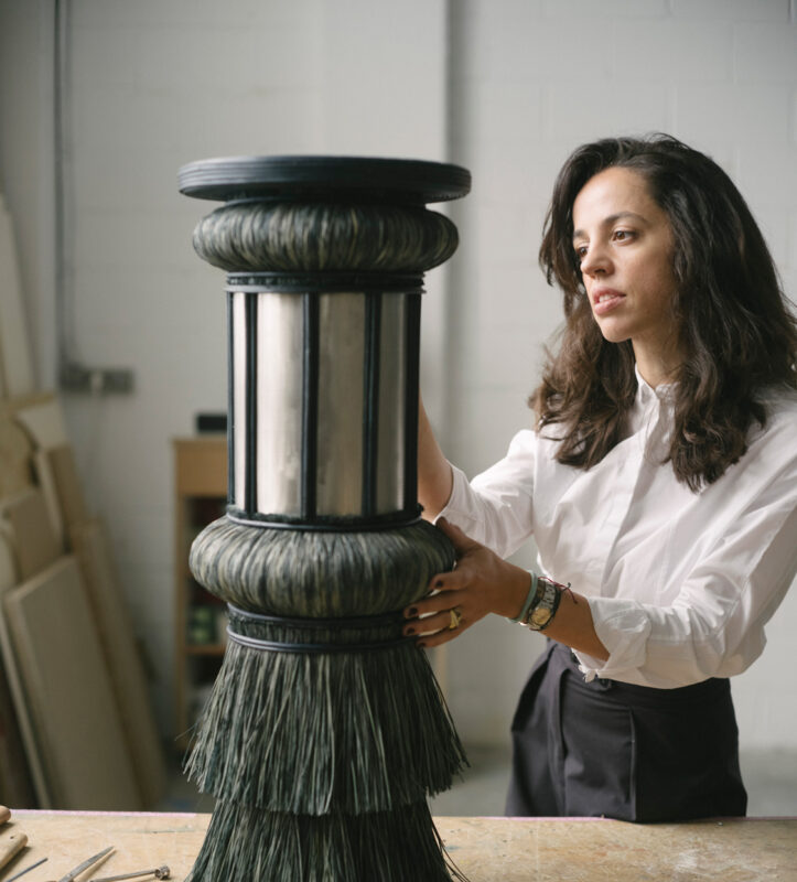 Woman in a white shirt adjusts a decorative column with a fringed base and mirrored section on a workbench.