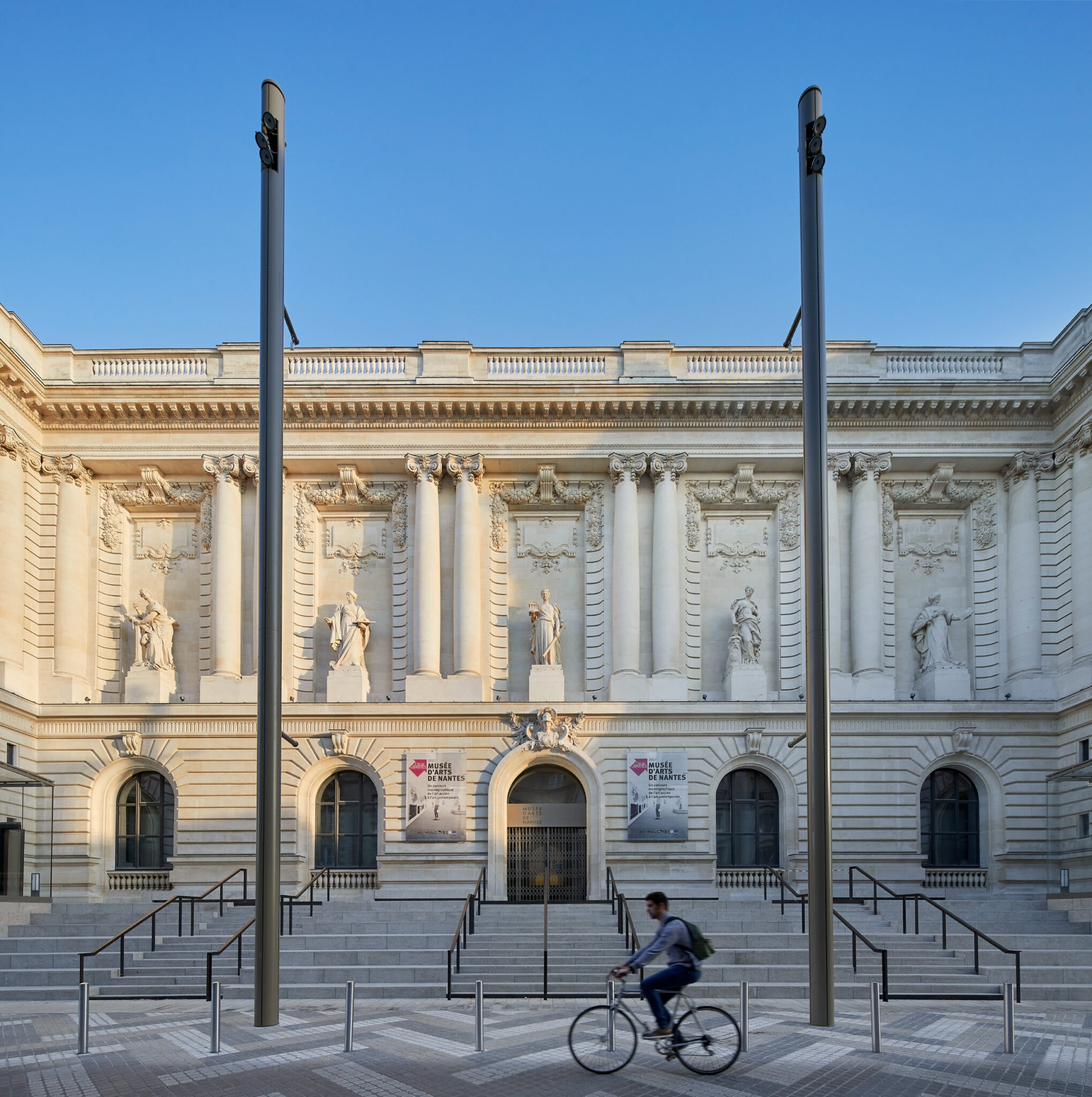 Historic architecture of the Musée d'Arts de Nantes with a cyclist passing by, under a clear blue sky.