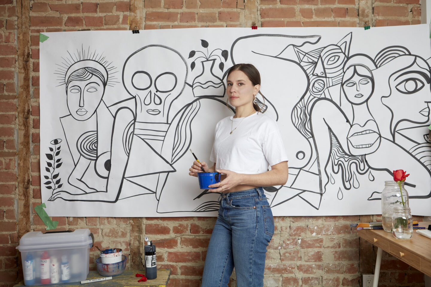 Artist holding a paintbrush and a blue container, standing in front of a large black-and-white mural in a brick studio.