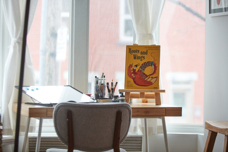 A wooden easel displays the book "Roots and Wings" by Hanna Barczyk, set on a desk with art supplies and natural light.