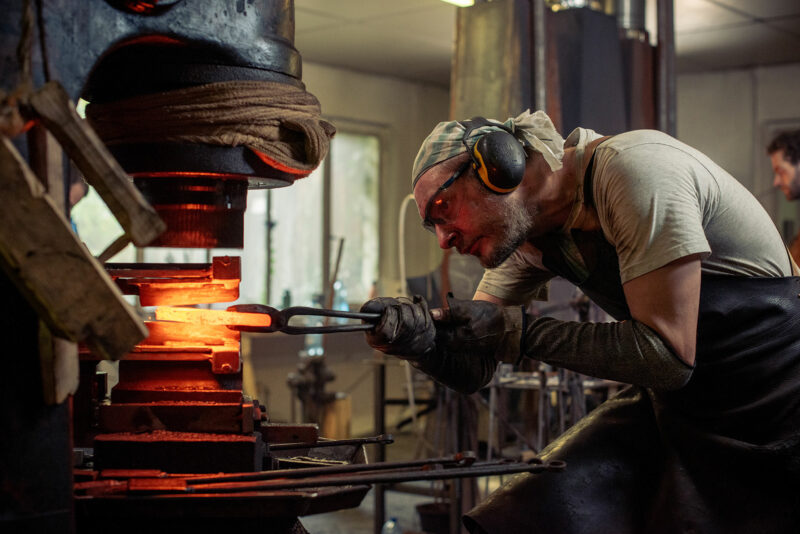 Blacksmith working with glowing metal in a workshop, wearing protective gear and focused on the task.