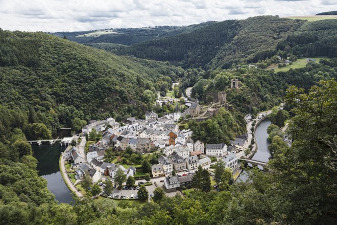 Aerial view of Esch-sur-Sûre, showcasing the village, river, and surrounding lush hills under a cloudy sky.