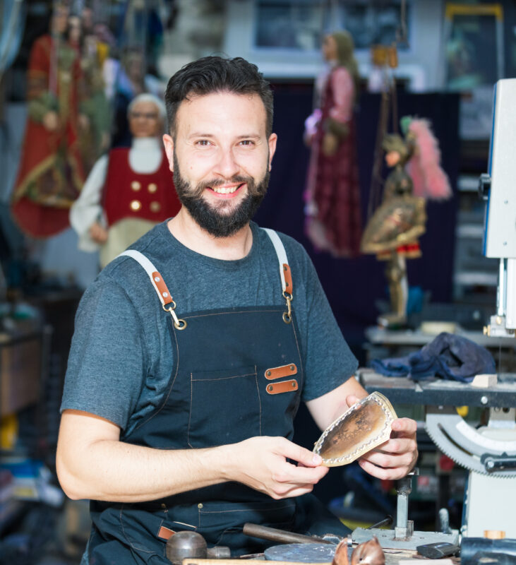 Man with a beard in a workshop, holding a crafted item, surrounded by puppets and tools.