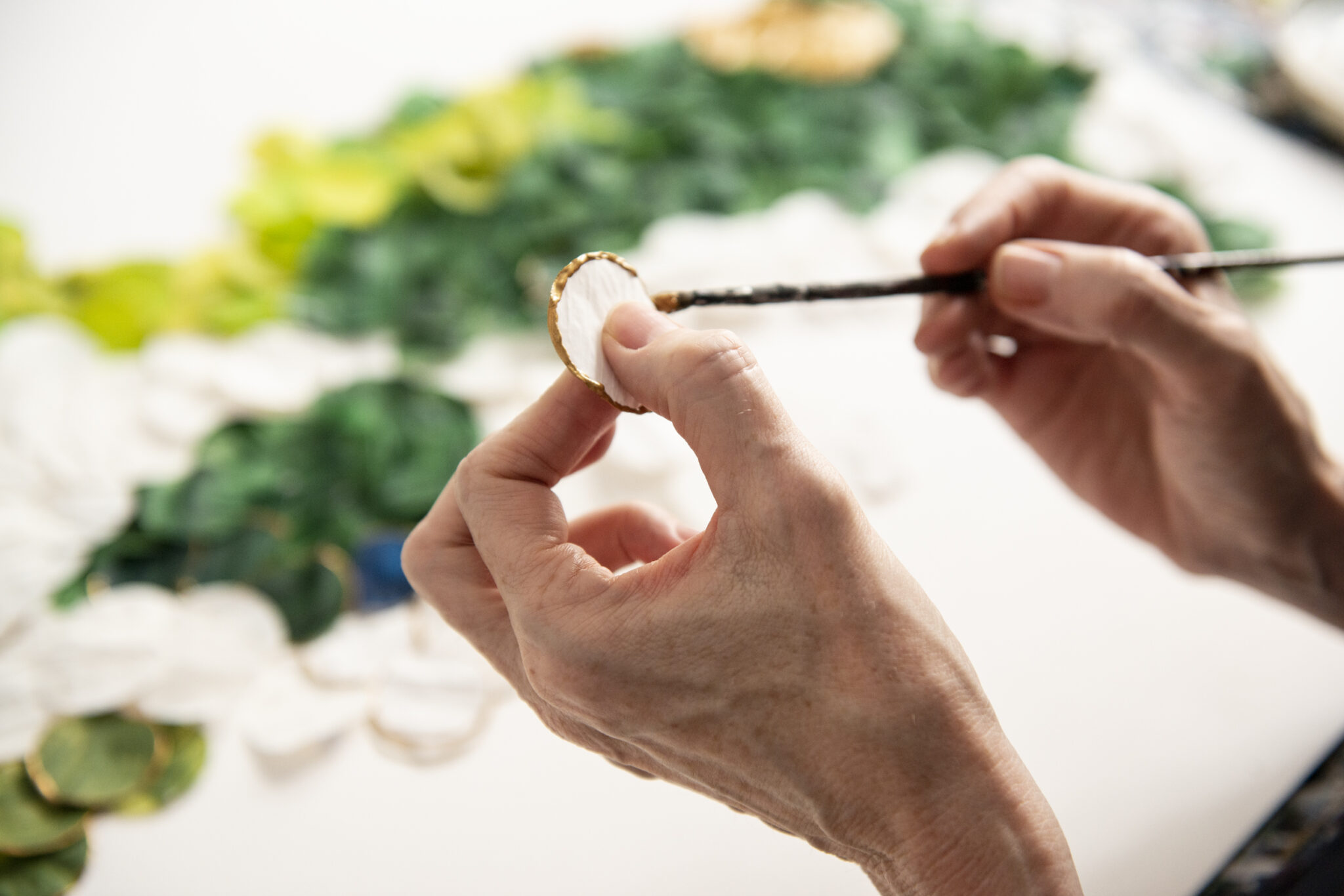 Hands holding a small circular object while painting, with a colorful background of green and white materials.