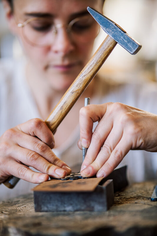 Jeweler Annie Sibert carefully hammers a metal piece while using a tool to engrave on a workbench.