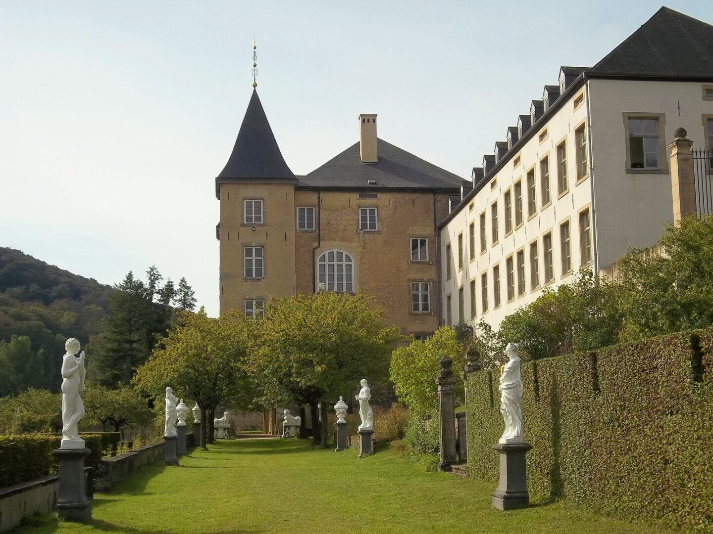 D’Ansembourg Castle gardens featuring classical statues, vibrant greenery, and the castle in the background.