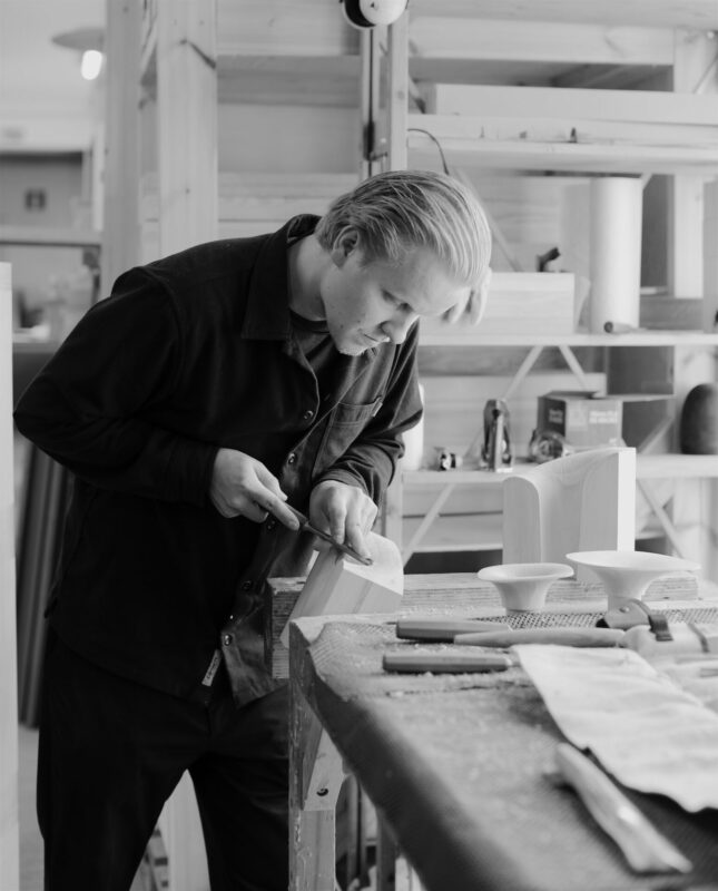 Craftsman shaping wood in a workshop, focused on his work, surrounded by tools and wooden pieces.