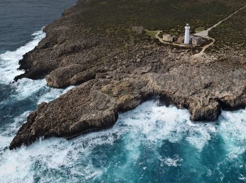 Waves crashing against rocky coastline near a lighthouse in Syracuse, with lush greenery in the background.