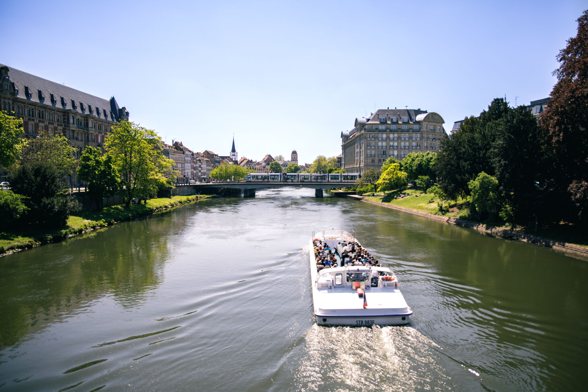 A boat with passengers navigates a calm river in Strasbourg, surrounded by greenery and historic buildings under a clear b...