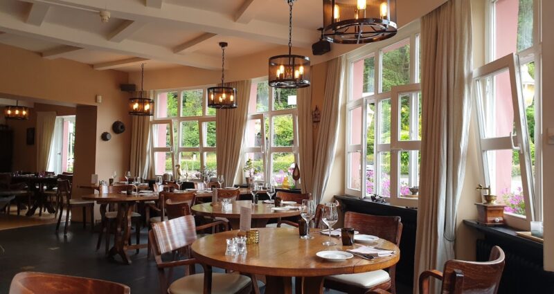 Rustic restaurant interior with wooden tables, elegant lighting, and large windows overlooking a garden.