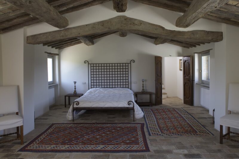 Spacious bedroom with a wooden beam ceiling, a bed with a white mattress, and decorative rugs on a stone floor.