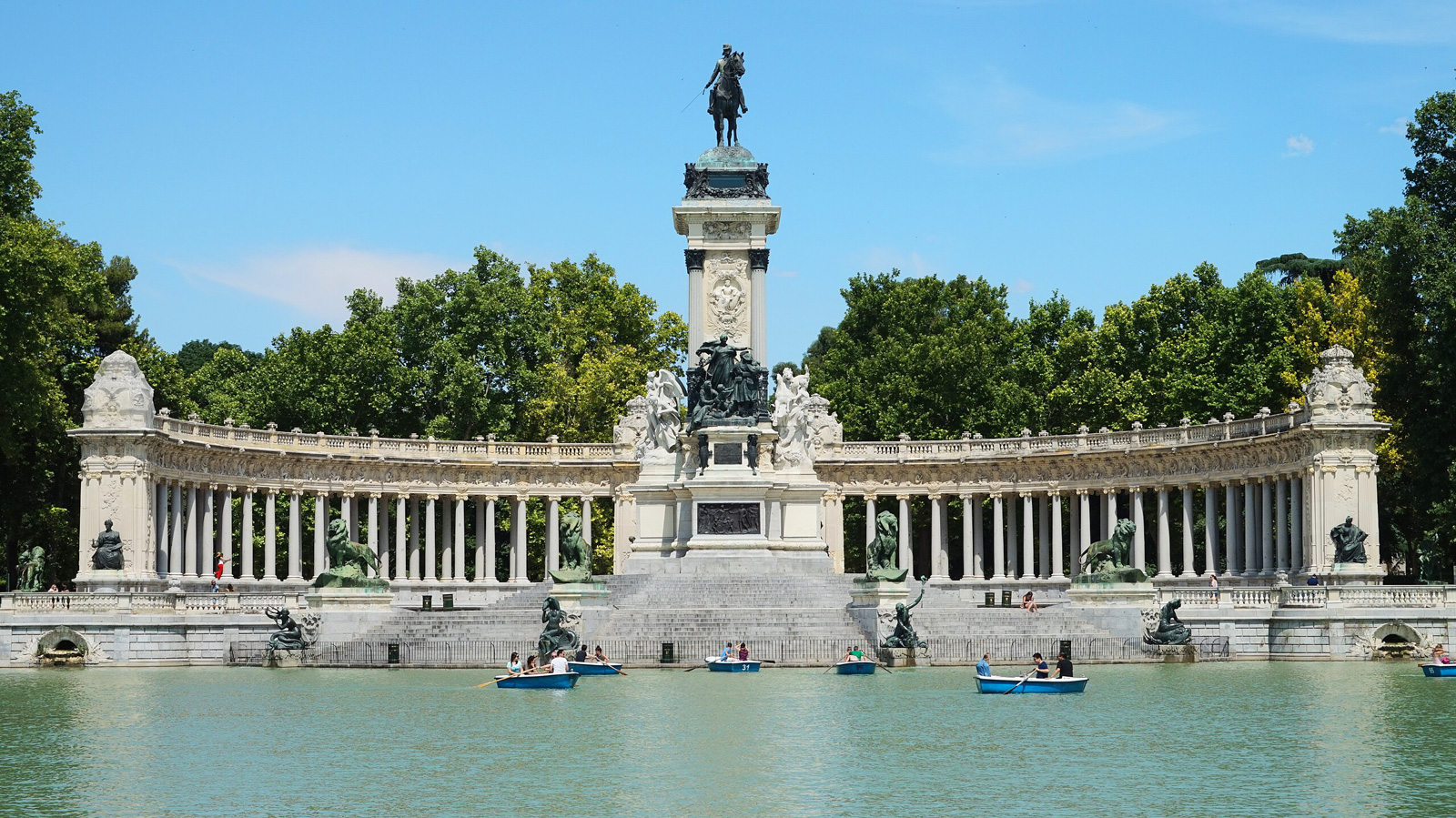 Monumental plaza with a statue atop a pedestal, surrounded by trees and a lake with small boats in Madrid.